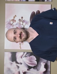 Smiling bald man with a mustache and beard wearing dark blue scrub top standing indoors in front of wall art featuring close-ups of flowers and stones.