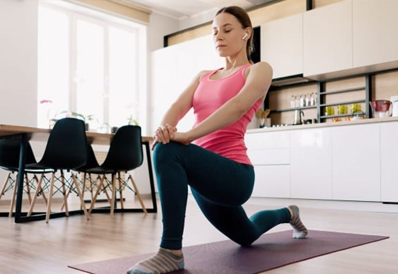 Woman in pink tank top and leggings doing a yoga lunge stretch on a mat in a bright kitchen.