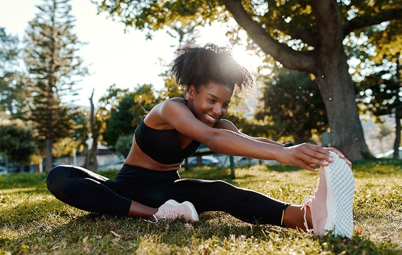 Woman in black workout clothes stretching leg forward on grass in a sunny park.