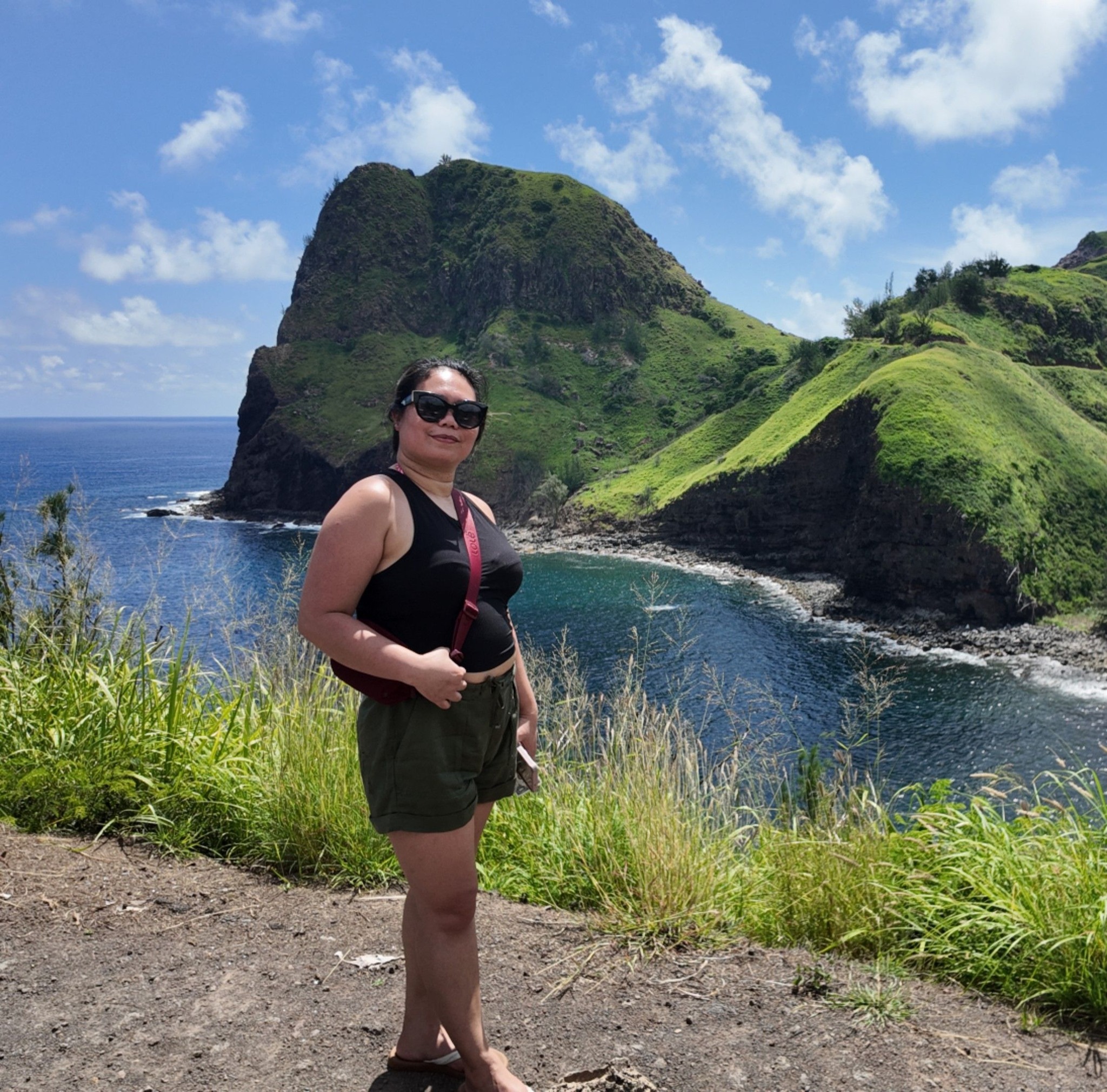 Woman in sunglasses posing near a grassy cliff with blue ocean and green hills in the background.