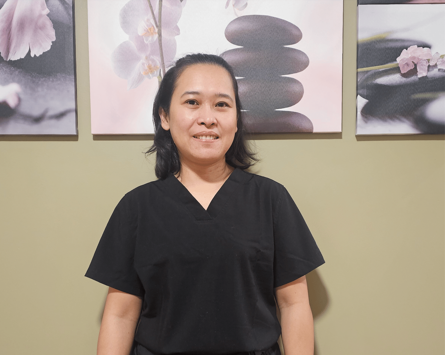 Smiling woman with shoulder-length hair wearing black scrubs stands in front of wall with three floral zen paintings.