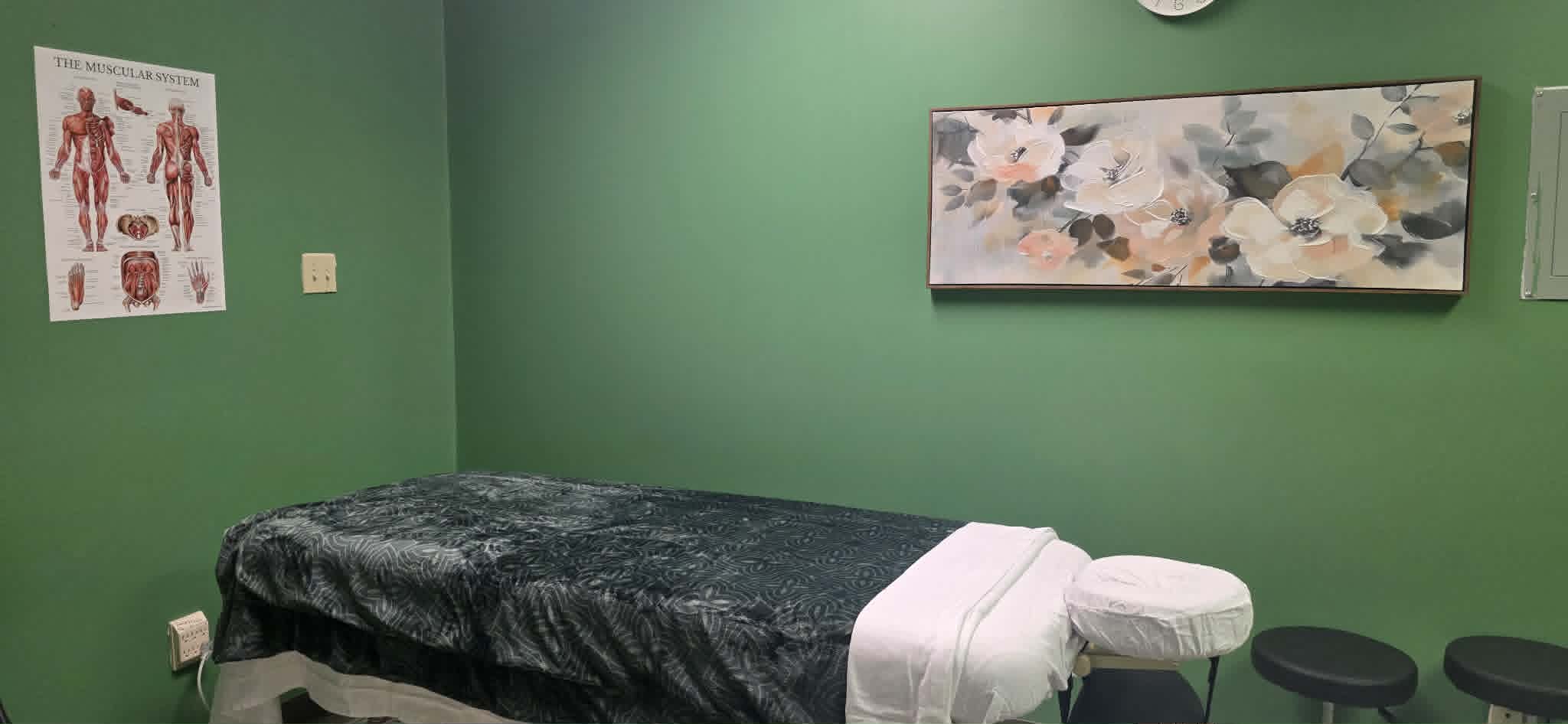 Massage table with dark cover and white towels in green-walled room with muscle chart and floral painting.